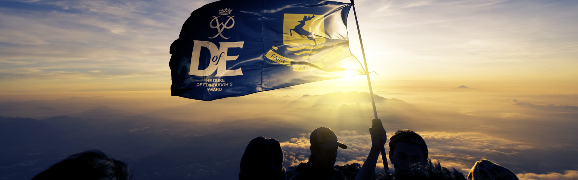 silouhetted figures hold up flag with school and duke of edinburgh logo at top of mountain